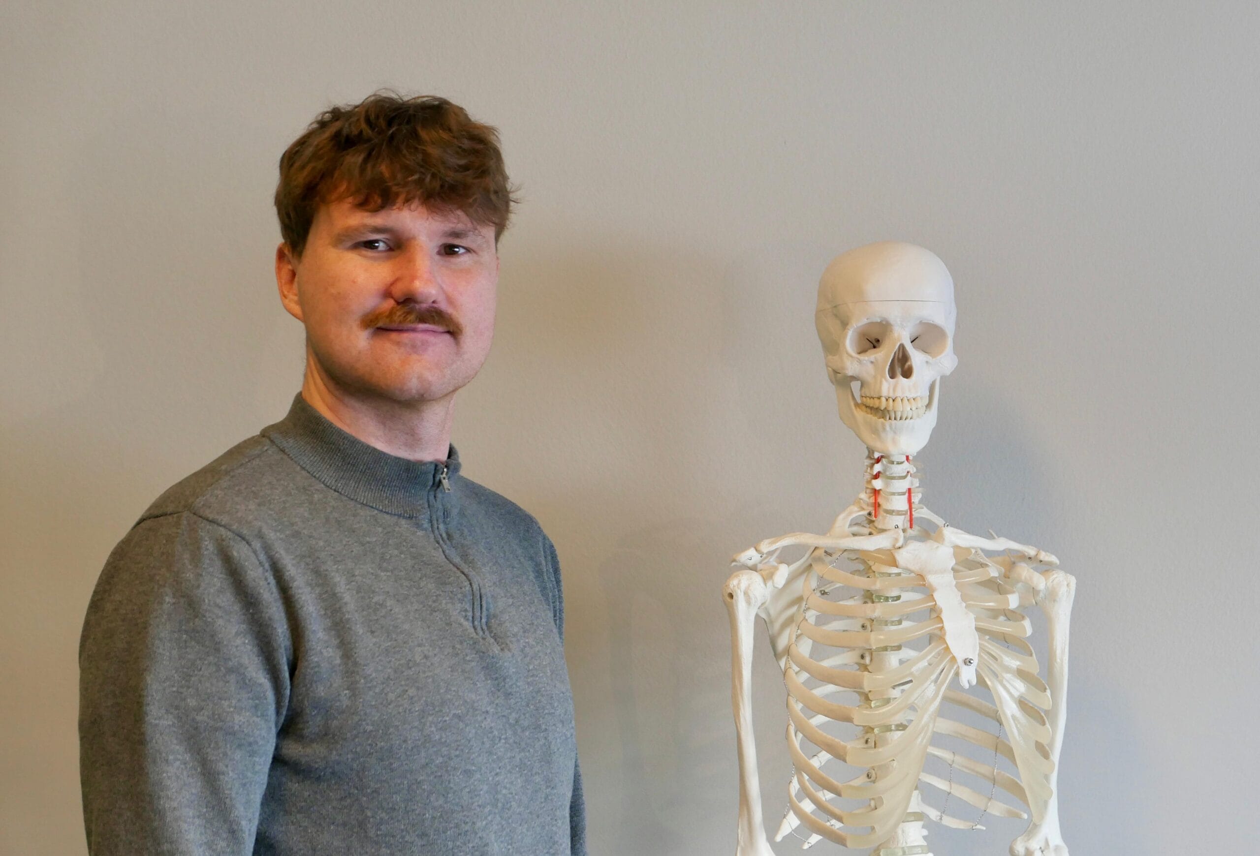 Therapeut steht neben einem Skelett in dem Praxisraum vor einer weißen Wand und lächelt in die Kamera