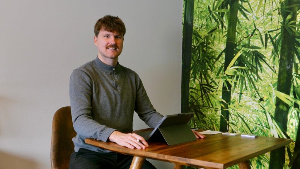 Empfang Freundlicher Empfangsbereich mit sitzender Person (Sebastian Ziegler) am Tisch und iPad vor einer grünen Pflanzenwandtapete