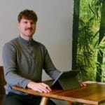 Empfang Freundlicher Empfangsbereich mit sitzender Person (Sebastian Ziegler) am Tisch und iPad vor einer grünen Pflanzenwandtapete