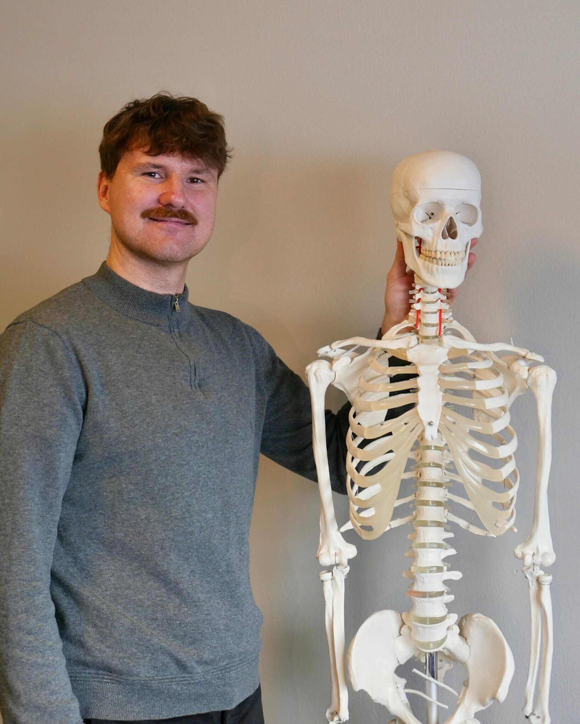 Therapeut steht neben einem Skelett Seine Hand liegt auf dem Skelett Er lächelt in die Kamera
