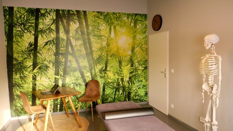 Praxisraum Behandlungsraum mit Therapieliege, Stuhl und beruhigenden Waldmotiv an der Wand in der Praxis von Sebastian Ziegler in München Bogenhausen. Nach Kontaktaufnahme und Terminvereinbarung werden Sie hier behandelt